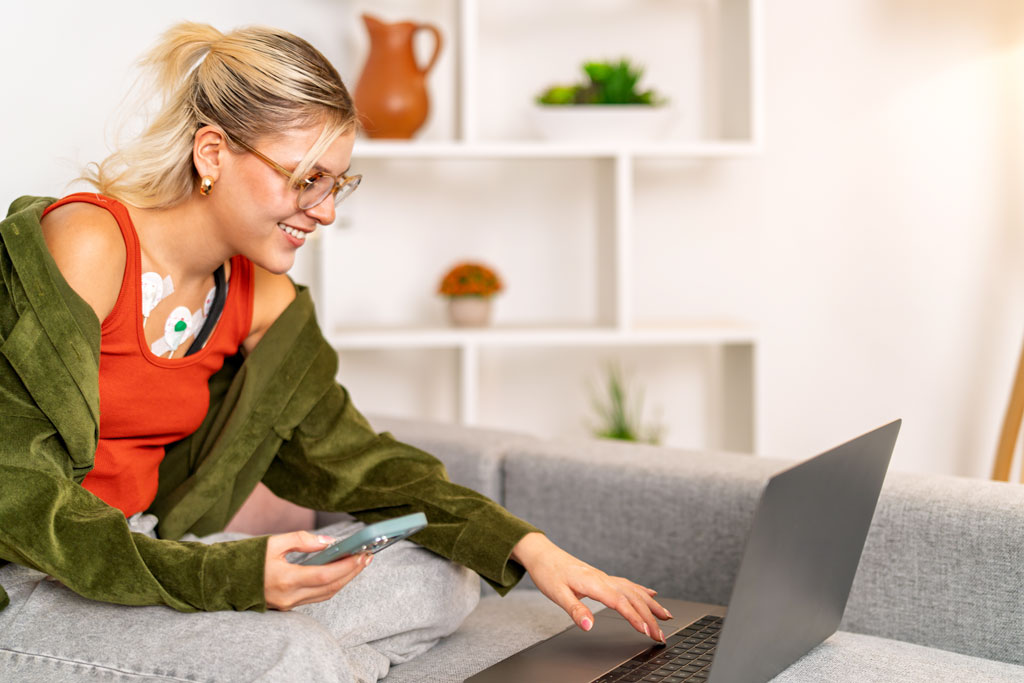 Patient wearing a holter monitor using a laptop and smartphone, transmitting her heart data to healthcare professional, enjoying comfort of home monitoring